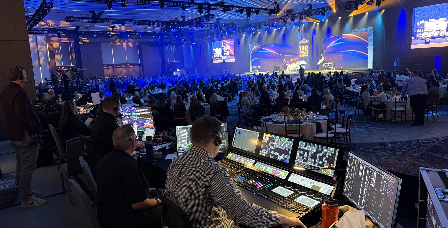 Three operators at a video village production desk at the back of a packed general session, with a wide blue-lit stage in front of them.