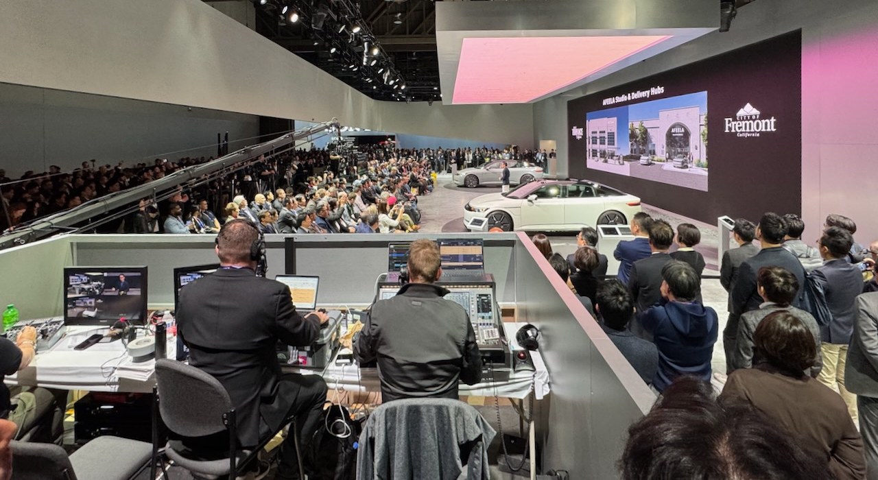 A front-of-house production desk with two operators in headsets overlooking a product launch stage, a packed press gallery and a crowd of attendees.