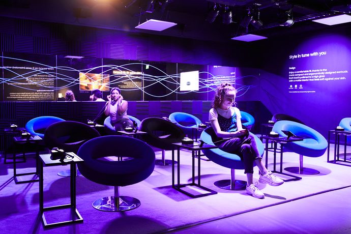 A talk-show style lounge set in deep purple light with two people seated on stage.