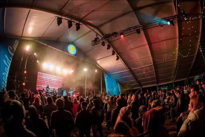 An outdoor festival stage at dusk under a curved canopy with a dense crowd in front.