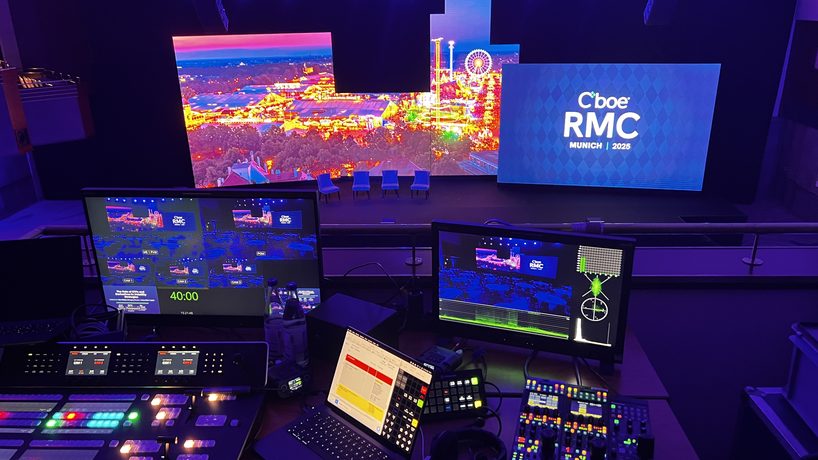 A broadcast-style news desk on a show floor with a camera framing two hosts.
